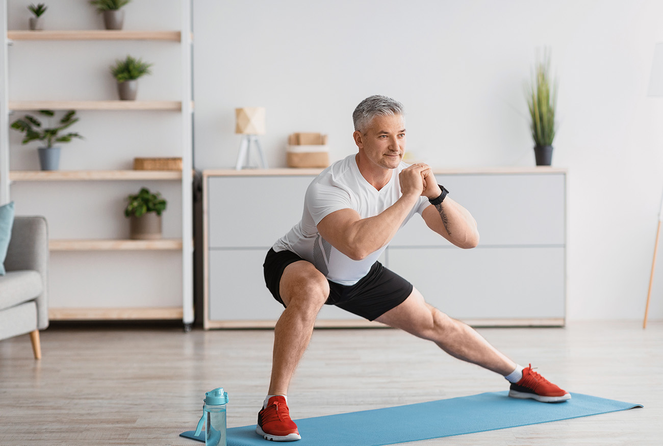 Home serobics. Mature man doing lunges exercises on mat in living room, enjoying domestic fitness training wearing sportswear and smiling, free space